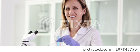 Happy laboratory worker holds tube with blue liquid at table Happy laboratory worker holds tube with blue liquid at table 107849759