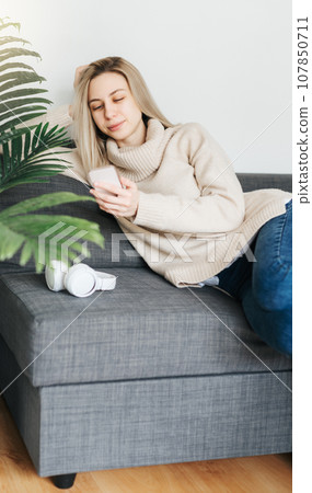 Happy young woman relaxing on comfortable couch, holding smartphone in hands. 107850711