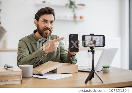 Young man recording video while unboxing smartphone indoors Young man recording video while unboxing smartphone indoors 107851294