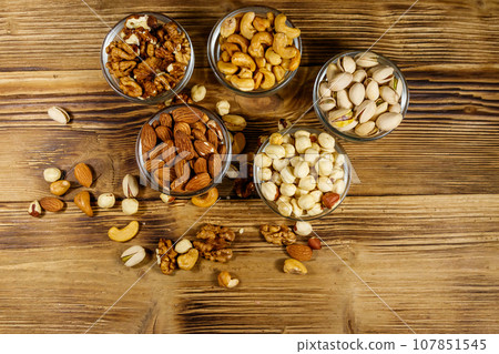 Assortment of nuts on wooden table. Almond, hazelnut, pistachio, walnut and cashew in glass bowls. Top view. Healthy eating concept Assortment of nuts on wooden table. Almond, hazelnut, pistachio, walnut and cashew in glass bowls. Top view. Healthy eating concept 107851545
