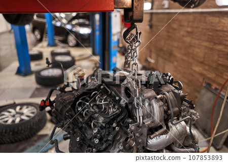 A car mechanic inspects the engine with a flashlight. Engine Block on a repair stand with Special crane jack. Technical service station for car. A car mechanic inspects the engine with a flashlight. Engine Block on a repair stand with Special crane jack. Technical service station for car. 107853893
