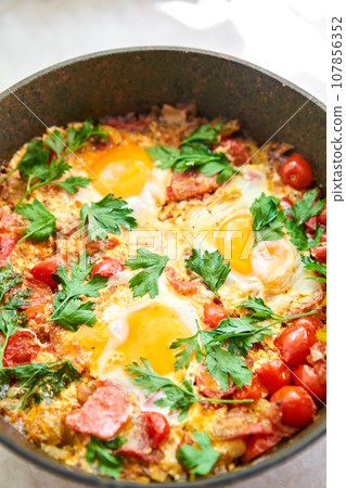 Shakshouka for breakfast. Homemade shakshuka, fried eggs, onion, bell pepper, tomatoes and parsley in a pan. Marble background. Top view. chakchouka Shakshouka for breakfast. Homemade shakshuka, fried eggs, onion, bell pepper, tomatoes and parsley in a pan. Marble background. Top view. chakchouka 107856352