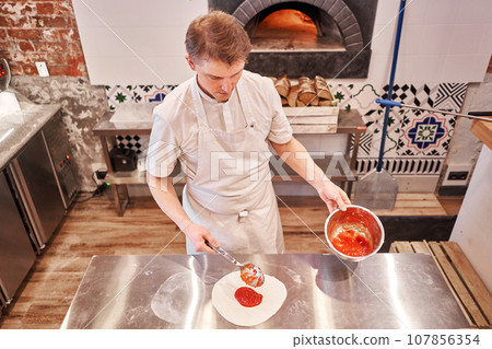 Cooking pizza. the workpiece poured tomato sauce. Closeup hand of chef baker in uniform apron cook at kitchen Cooking pizza. the workpiece poured tomato sauce. Closeup hand of chef baker in uniform apron cook at kitchen 107856354