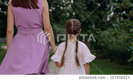Mother and little daughter walk joining hands along road across city park Mother and little daughter walk joining hands along road across city park 107856825