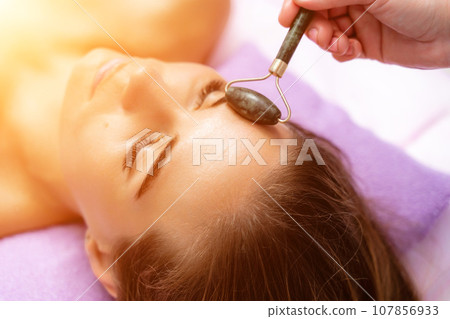 Facial massage. Side view european woman getting massage with jade face roller gouache in spa salon Facial massage. Side view european woman getting massage with jade face roller gouache in spa salon 107856933