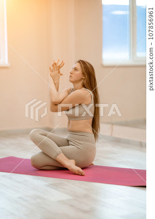 Young woman doing yoga in the gym. A girl with long hair and in a beige tracksuit stands in a cow pose on a pink carpet. A woman performs Gomukhasana. 107856961