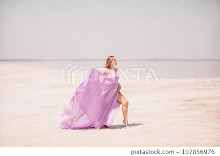 Woman pink salt lake. Against the backdrop of a pink salt lake, a woman in a long pink dress takes a leisurely stroll along the white, salty shore, capturing a wanderlust moment. 107856976