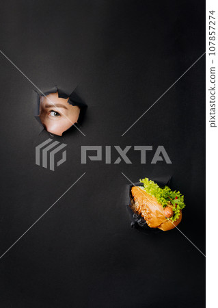 Female hand holding hot dog and looks through holes in black paper background. Street food concept Female hand holding hot dog and looks through holes in black paper background. Street food concept 107857274