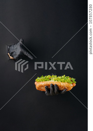 Hand holding hot dog and and point finger on it through torn holes in black paper background. Street food concept Hand holding hot dog and and point finger on it through torn holes in black paper background. Street food concept 107857280
