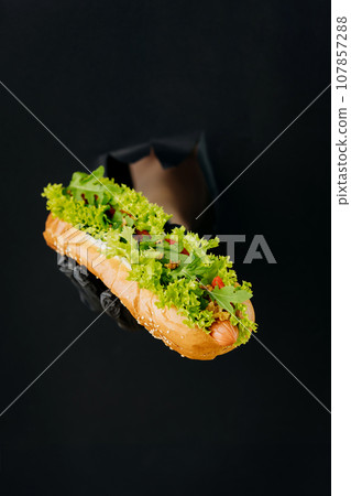 Hand holding hot dog through torn holes in black paper background. Street food concept Hand holding hot dog through torn holes in black paper background. Street food concept 107857288