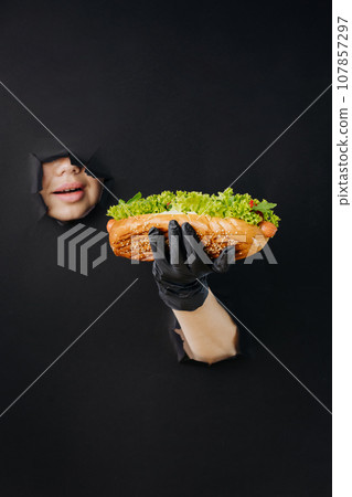 Female hand holding hot dog and looks through holes in black paper background. Street food concept 107857297