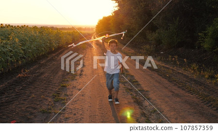 Happy little boy runs playing with kite along rural road at sunset light Happy little boy runs playing with kite along rural road at sunset light 107857359