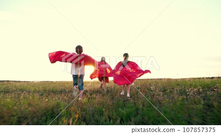 Happy children with red capes play heroes running across grassy field Happy children with red capes play heroes running across grassy field 107857375