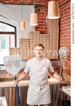 The chef prepares pizza in a neapolitan pizzeria. Cook in a apron in the kitchen with a shovel in his hands. boxes for food delivery on background. The chef prepares pizza in a neapolitan pizzeria. Cook in a apron in the kitchen with a shovel in his hands. boxes for food delivery on background. 107857515
