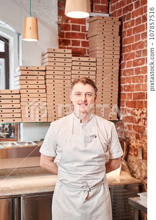 The chef prepares pizza in a neapolitan pizzeria. Cook in a apron in the kitchen with a shovel in his hands. boxes for food delivery on background. The chef prepares pizza in a neapolitan pizzeria. Cook in a apron in the kitchen with a shovel in his hands. boxes for food delivery on background. 107857516