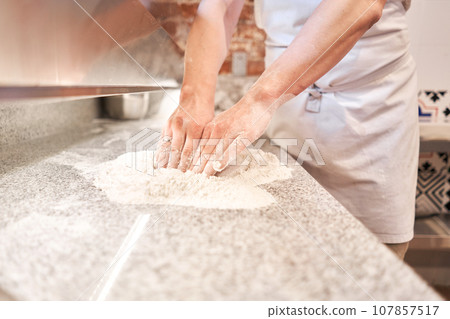 The chef prepare pizza. Raw pizza ready to bake. Cook in a apron in the kitchen Rolls out pizza dough. boxes for food delivery on background. The chef prepare pizza. Raw pizza ready to bake. Cook in a apron in the kitchen Rolls out pizza dough. boxes for food delivery on background. 107857517