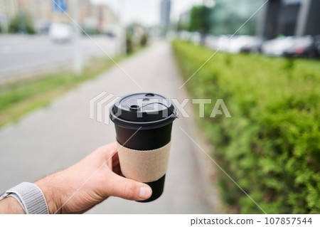 A cup of hot drink in the hands of a man. A man in the park holds a cup of coffee in her hands. Breakfast on the go. The concept of a picnic and going out. 107857544