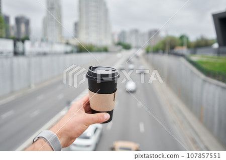 A cup of hot drink in the hands of a man. A man on the city highway holds a cup of coffee in her hands. Breakfast on the go. The concept of a picnic and going out. 107857551