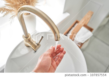 A man's hand touches the water in a beautiful sink with a golden faucet next to an oval mirror and a shelf with hand towels. Close-up of an elegant golden faucet in the bathroom sink next to stylish 107857554