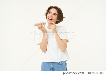 Portrait of brunette woman recording voice message, girl talking on mobile phone, using speakerphone, translating her words, standing over white background Portrait of brunette woman recording voice message, girl talking on mobile phone, using speakerphone, translating her words, standing over white background 107860108