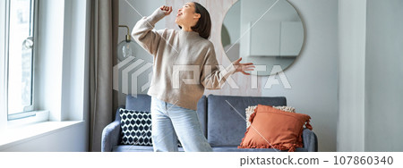 People and emotions. Portrait of asian girl standing in living room, singing in shadow microphone, dancing and having fun, feeling joy 107860340