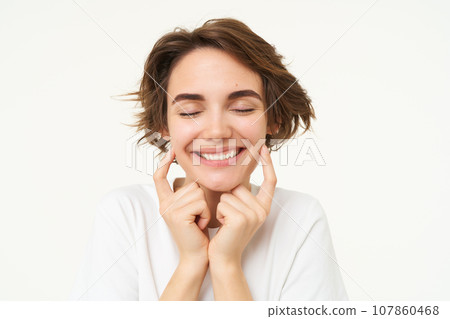 Portrait of brunette woman smiling, shuts her eyes and touches dimples on her cheeks, stands over white background Portrait of brunette woman smiling, shuts her eyes and touches dimples on her cheeks, stands over white background 107860468