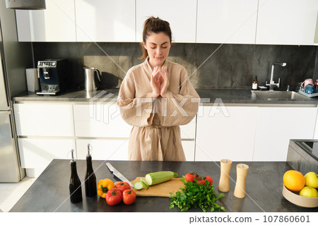 Portrait of excited woman looks thoughtful at chopping board with zucchini, vegetables, preparing healthy salad, making vegetarian meal, choosing what to have for dinner, standing in kitchen Portrait of excited woman looks thoughtful at chopping board with zucchini, vegetables, preparing healthy salad, making vegetarian meal, choosing what to have for dinner, standing in kitchen 107860601