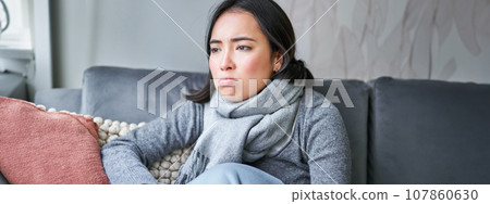 Portrait of sad woman feeling unwell, wearing warm clothes and scarf at her home without heating, staying on sick leave in her house Portrait of sad woman feeling unwell, wearing warm clothes and scarf at her home without heating, staying on sick leave in her house 107860630