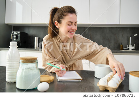Attractive young cheerful girl baking at the kitchen, making dough, holding recipe book, having ideas 107860723
