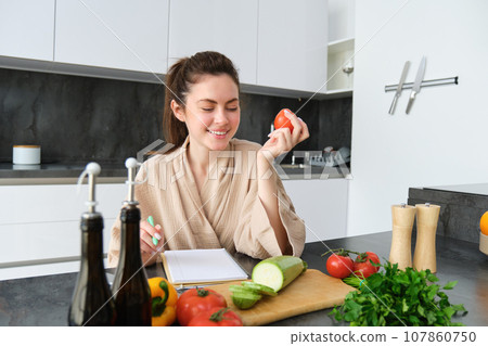 Portrait of beautiful young woman in the kitchen, writing down cooking recipe, sitting near chopping board with vegetables and making grocery list, creating healthy veggie menu for her family 107860750