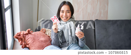 Portrait of happy smiling asian girl showing medication, sore throat spray and drugs from flu or cold, staying at home, wearing warm clothes Portrait of happy smiling asian girl showing medication, sore throat spray and drugs from flu or cold, staying at home, wearing warm clothes 107860815