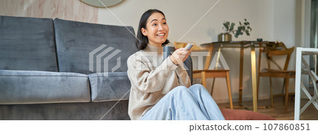 Portrait of young korean woman watching television, holding remote and looking amazed at tv screen, spending time at home Portrait of young korean woman watching television, holding remote and looking amazed at tv screen, spending time at home 107860851