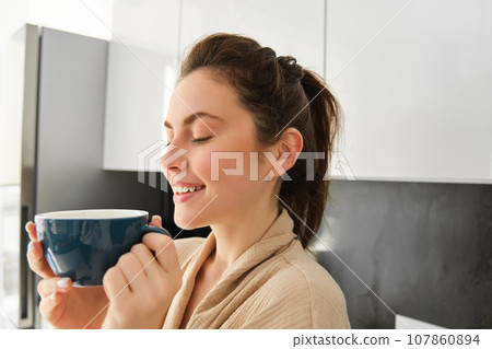 Portrait of happy young woman starts her morning with mug of coffee, drinking tea from cup, standing in the kitchen, smiling cheerfully 107860894