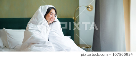 Cozy mornings. Happy asian girl feels warm in her bed, covers herself with cozy duvet in her bedroom, a comfortable stay in hotel room 107860954