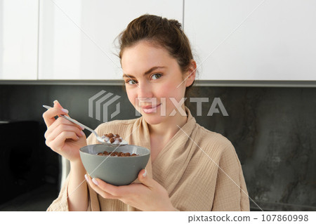 Good-looking brunette woman with bowl of cereals, eating meal for breakfast, wearing bathrobe, looking at camera and smiling Good-looking brunette woman with bowl of cereals, eating meal for breakfast, wearing bathrobe, looking at camera and smiling 107860998