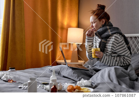 ill female has bad cough, sitting on bed at home, drinking hot tea and working on laptop ill female has bad cough, sitting on bed at home, drinking hot tea and working on laptop 107861087
