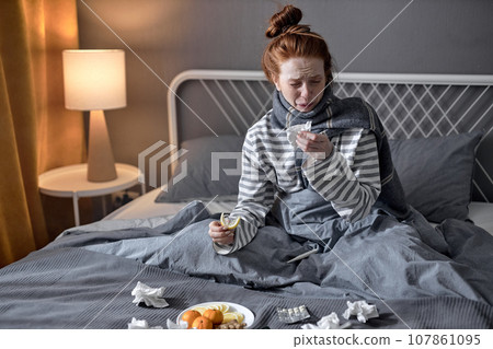 young woman with cold blowing runny nose. female sneezing in tissue in bedroom 107861095