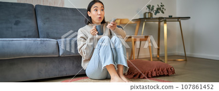 Portrait of girl watching television at home, sits on floor near sofa, holds remote and changes channels Portrait of girl watching television at home, sits on floor near sofa, holds remote and changes channels 107861452