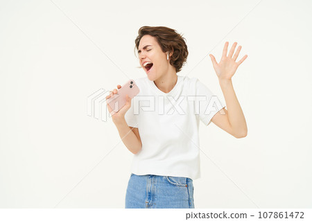 Portrait of happy young woman singing in mobile speakerphone, listening music in wireless headphonse, playing smartphone karaoke game, standing over white background Portrait of happy young woman singing in mobile speakerphone, listening music in wireless headphonse, playing smartphone karaoke game, standing over white background 107861472