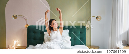 Portrait of smiling happy asian girl, wakes up feeling enthusiastic, stretches her hands up, enjoys good morning in her bedroom Portrait of smiling happy asian girl, wakes up feeling enthusiastic, stretches her hands up, enjoys good morning in her bedroom 107861509