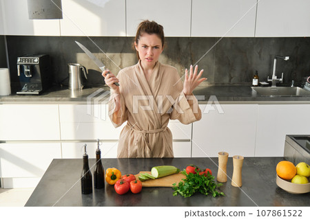 Portrait of annoyed woman with knife, angry while cooking in the kitchen, frustrated while doing house chores and preparing food for family, standing in bathrobe 107861522