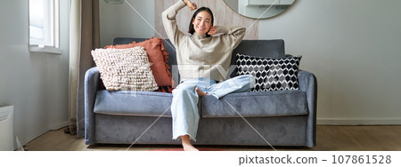 Portrait of happy asian woman feeling lazy, stretching on sofa and smiling pleased, relaxing at home, resting from work Portrait of happy asian woman feeling lazy, stretching on sofa and smiling pleased, relaxing at home, resting from work 107861528