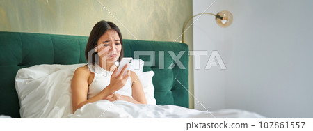 Portrait of concerned asian woman holding mobile phone, receive bad phone call, looking worried and upset, having difficult telephone conversation while lying in bed Portrait of concerned asian woman holding mobile phone, receive bad phone call, looking worried and upset, having difficult telephone conversation while lying in bed 107861557