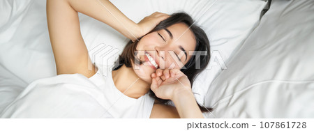 Close up of happy asian girl lying in bed on her pillow, smiling and rolling over as feeling energetic in morning, waking up early 107861728