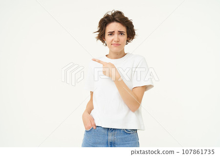Portrait of sceptical young woman, pointing finger left and looking with doubt at camera, dislike smth, standing over white studio background Portrait of sceptical young woman, pointing finger left and looking with doubt at camera, dislike smth, standing over white studio background 107861735