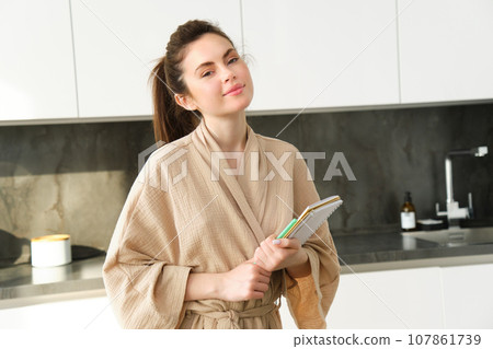 Portrait of beautiful modern woman, writing down grocery list, meal ideas in notebook, standing in the kitchen, wearing cosy bathrobe Portrait of beautiful modern woman, writing down grocery list, meal ideas in notebook, standing in the kitchen, wearing cosy bathrobe 107861739