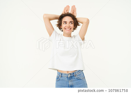 Portrait of funny and cute young woman with bunny ears gesture, holding palms on top of her head, smiling and looking happy at camera, standing over white background Portrait of funny and cute young woman with bunny ears gesture, holding palms on top of her head, smiling and looking happy at camera, standing over white background 107861758