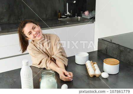 Attractive young cheerful girl baking at the kitchen, making dough, holding recipe book, having ideas Attractive young cheerful girl baking at the kitchen, making dough, holding recipe book, having ideas 107861779