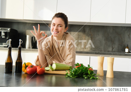 Portrait of smiling brunette girl making vegan dinner, showing okay sign, recommending healthy food, chopping vegetables in kitchen for salad or for vegan breakfast Portrait of smiling brunette girl making vegan dinner, showing okay sign, recommending healthy food, chopping vegetables in kitchen for salad or for vegan breakfast 107861781