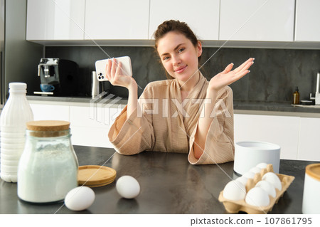 Attractive young cheerful girl baking at the kitchen, making dough, holding recipe book, having ideas 107861795